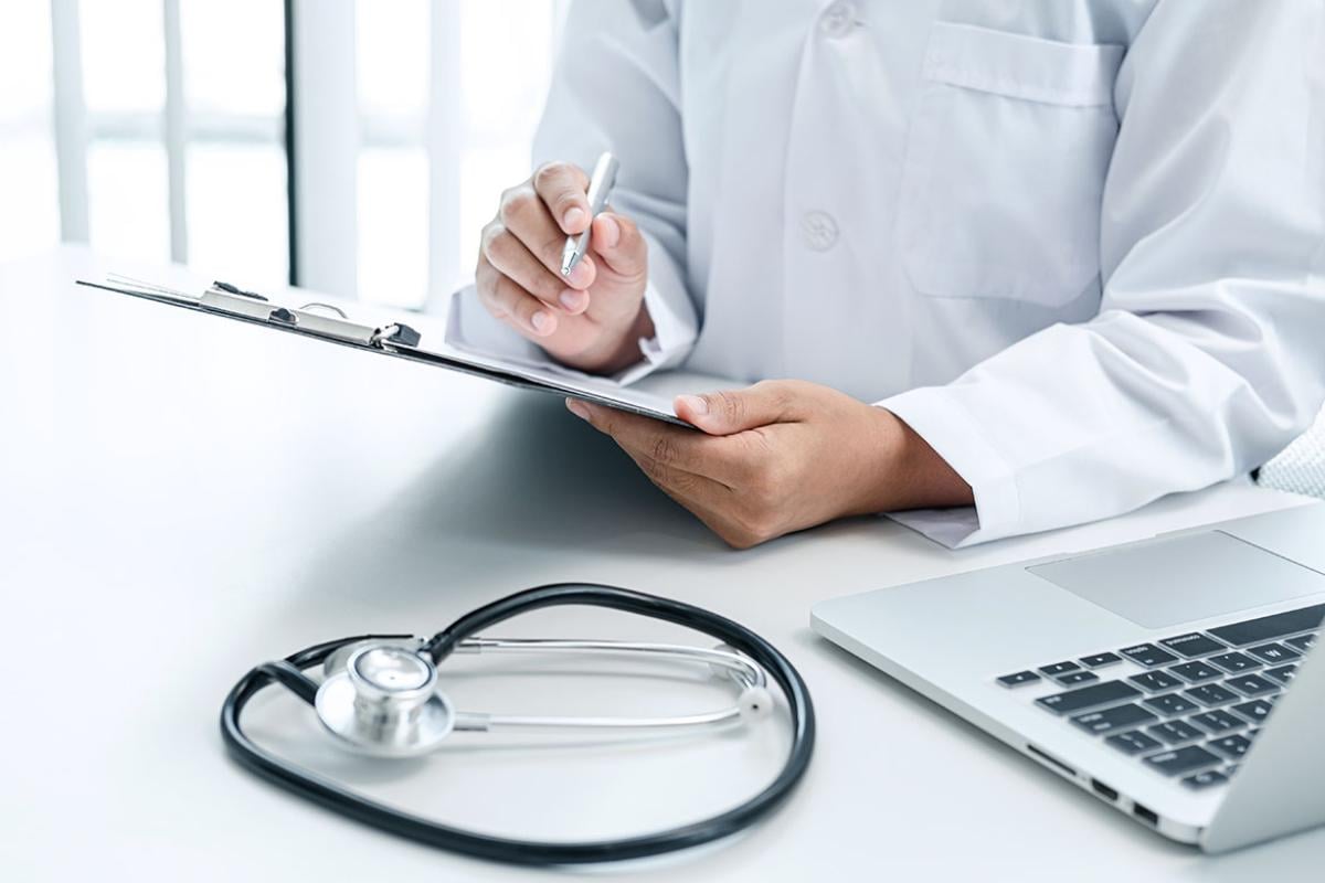 Person holding a clipboard with a laptop and stethoscope nearby
