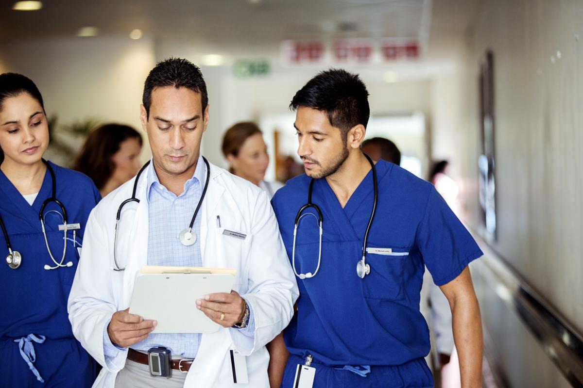 One female and two male residents walk down a hospital hallway examining a patient's health records.