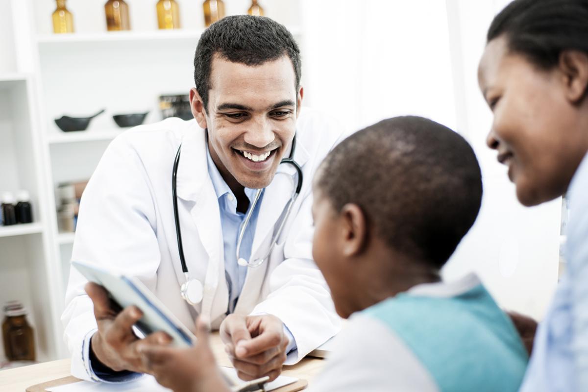 A young boy sits on his mother's lap and looks at the screen of his smiling physician's tablet.